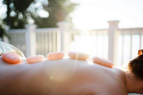 A person lying on their stomach outdoors with smooth stones placed along their back, sunlight shining in the background.