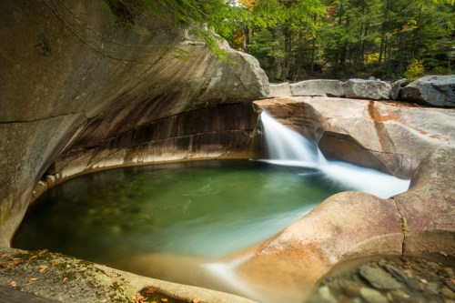 A serene waterfall cascades into a smooth, circular rock pool surrounded by lush greenery and rugged stone formations.