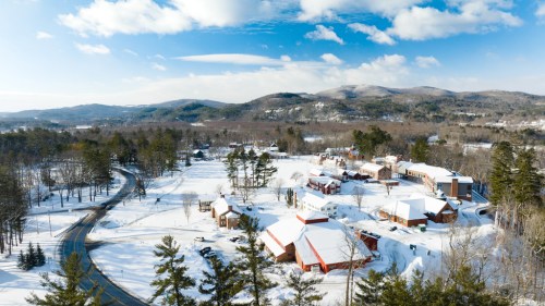Aerial view of a snowy landscape with a small village, surrounded by mountains and trees under a clear blue sky.