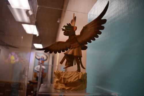 A wooden sculpture of a figure with wings, standing on a rocky base, displayed in a glass case.