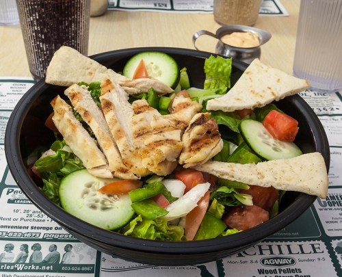 A fresh salad with grilled chicken, cucumbers, tomatoes, and pita bread, served in a black bowl on a table.
