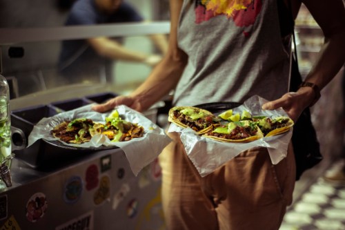 A person holds two plates of tacos topped with meat, onions, and lime, in a casual dining setting.
