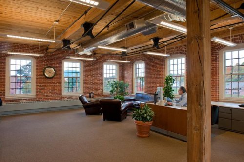 Modern office space with exposed brick walls, wooden beams, and a cozy seating area. A person works at a desk.