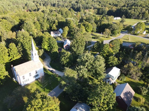 Aerial view of a small town with a church, houses, and lush green trees surrounding a winding road.