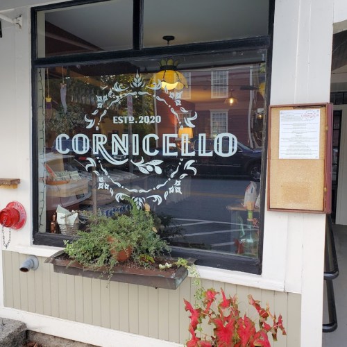 Window of a restaurant named "Cornicello," established in 2020, with plants and a menu board nearby.