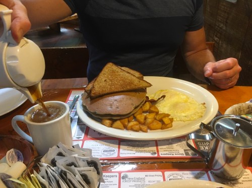 A person pours coffee next to a plate of a sandwich, eggs, and potatoes at a diner table.