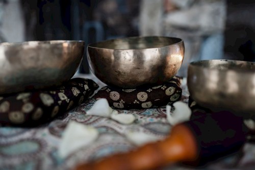 Three metal singing bowls on patterned fabric, surrounded by rose petals and a wooden mallet.