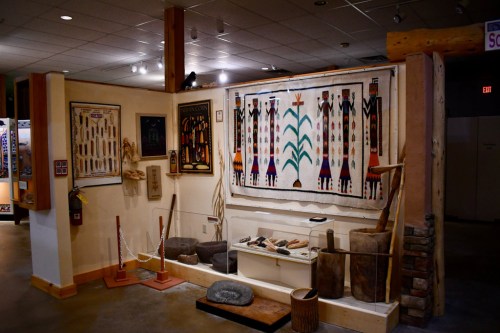 A museum display featuring Native American artifacts, including textiles, tools, and wall art in a well-lit room.
