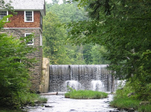 A stone building beside a waterfall, surrounded by lush greenery and trees. Water cascades over the dam into a stream.
