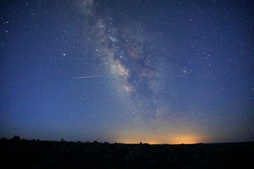 A stunning night sky filled with stars and the Milky Way, with a faint glow on the horizon and a shooting star.