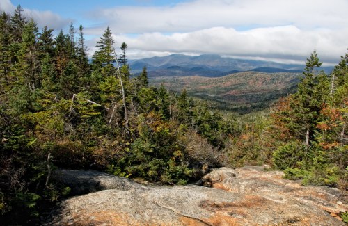 A scenic view of mountains and forests under a partly cloudy sky, showcasing vibrant autumn foliage.