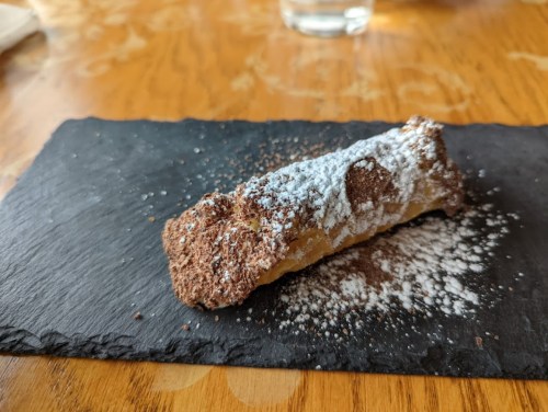 A single cannoli dusted with powdered sugar, resting on a black slate plate.