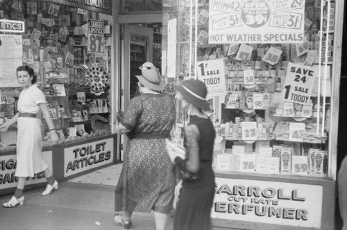 Black and white photo of women walking past a shop window filled with advertisements and various products.