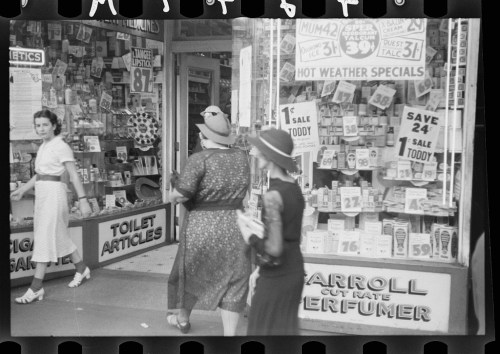 Two women walk past a storefront filled with vintage advertisements and signs for various products and sales.