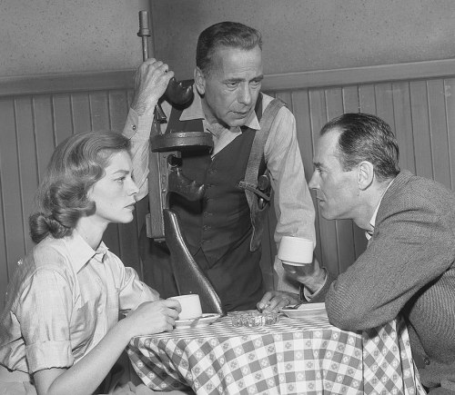 A tense conversation at a table between a woman and two men, one holding a phone, in a vintage setting.