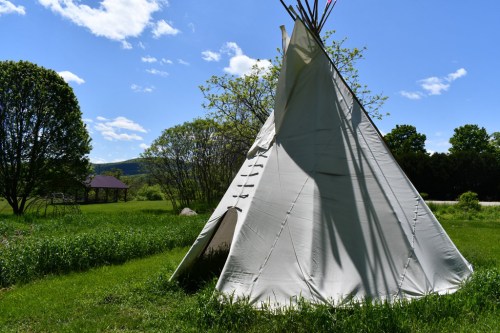 A white teepee stands in a grassy field under a blue sky with fluffy clouds, surrounded by trees and distant hills.