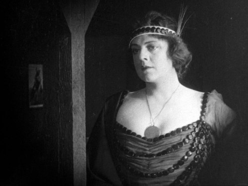 A woman in vintage attire with a headband and necklace, standing in a dimly lit room, looking thoughtfully to the side.