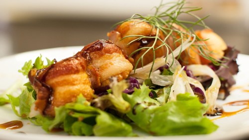 Plated dish featuring crispy bacon-wrapped bites on a bed of mixed greens and drizzled with sauce.