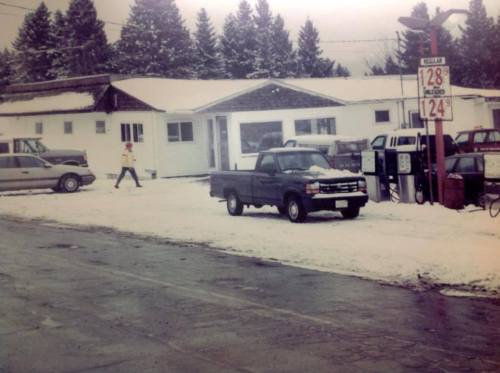 A snowy scene featuring a gas station, parked trucks, and a person walking by in winter attire. Pine trees in the background.