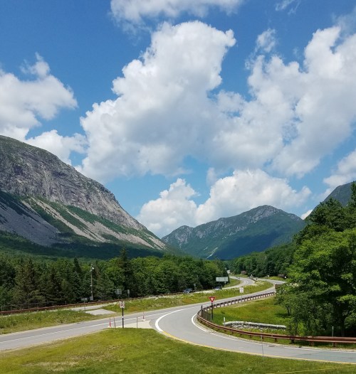 A winding road curves through lush green hills and mountains under a bright blue sky with fluffy white clouds.