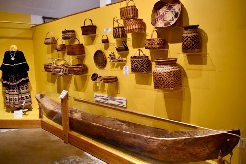 A display of traditional woven baskets and a wooden canoe against a yellow wall in a museum setting.