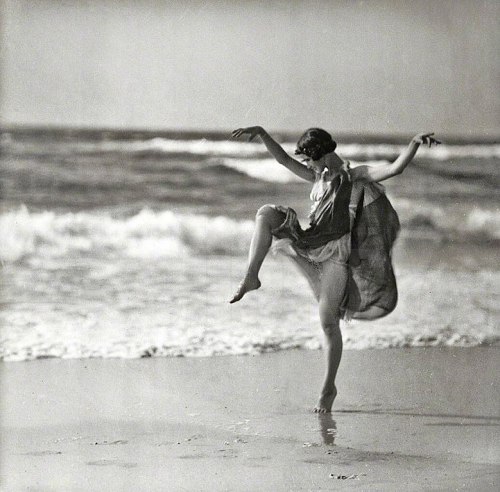 A woman in a vintage dress dances playfully by the ocean, with waves gently crashing in the background.