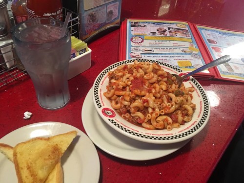 A plate of pasta with meat and vegetables, a glass of water, and a slice of toast on a red table. Menu in the background.
