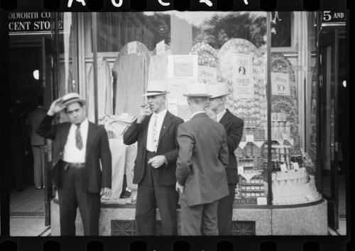Four men in hats stand outside a store, with a display of goods visible in the window behind them.