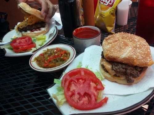 Two plates with burgers, one partially assembled, alongside sliced tomatoes, lettuce, and a bowl of salsa.