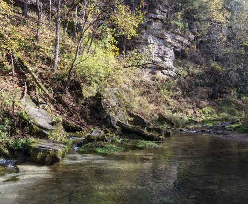 A serene riverbank scene with rocky cliffs, lush greenery, and clear water reflecting autumn foliage.