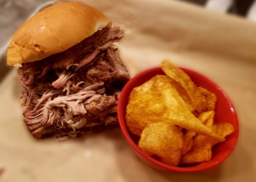 A pulled pork sandwich on a bun next to a bowl of crispy potato chips on a parchment-lined surface.