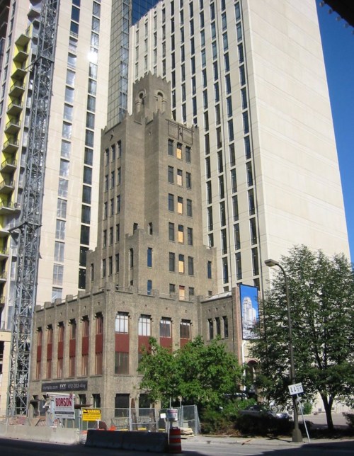 Historic building with a tall, narrow design, surrounded by modern skyscrapers and greenery.