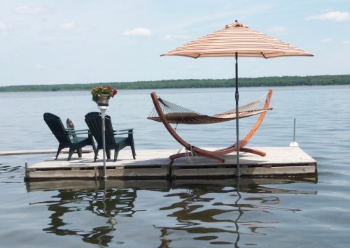 A serene lakeside scene featuring a hammock, umbrella, and chairs on a wooden dock, reflecting in calm water.