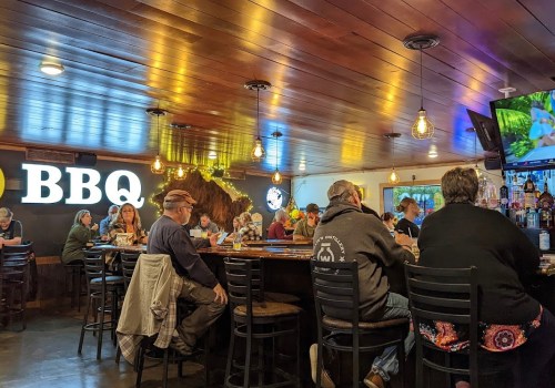 A lively BBQ restaurant interior with patrons seated at the bar and tables, warm lighting, and a large BBQ sign.