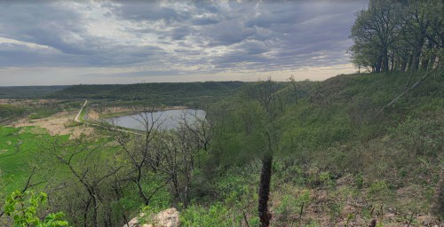 A panoramic view of a lush green landscape with a lake, under a cloudy sky, surrounded by trees and hills.