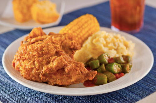A plate of fried chicken with corn, mashed potatoes, and okra, served on a blue tablecloth with a drink in the background.
