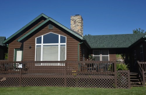 A brown log cabin with a stone chimney, large windows, and a wooden deck surrounded by green grass.