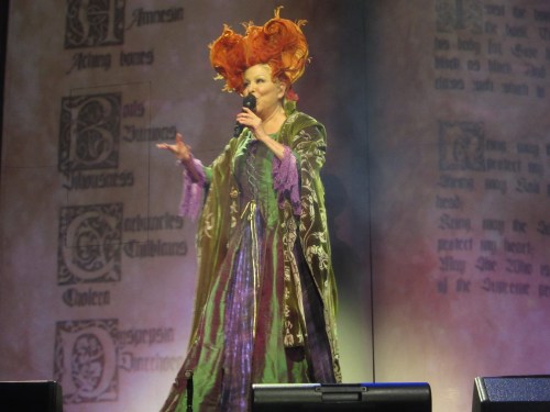 A performer in a vibrant, elaborate costume with orange hair gestures while singing on stage.