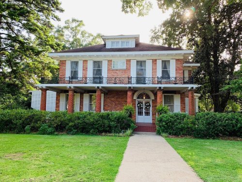 A large, two-story brick house with a porch, surrounded by greenery and trees, under a sunny sky.