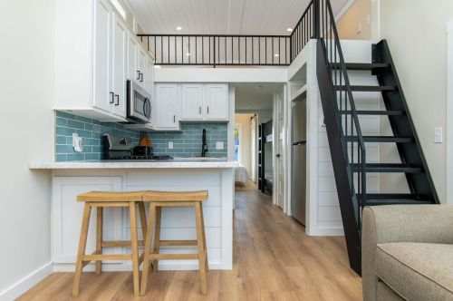 Modern kitchen with white cabinets, blue backsplash, and wooden stools, leading to a loft and staircase in a cozy interior.