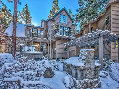 A cozy mountain home surrounded by snow-covered trees and a stone patio, featuring a pergola and clear blue sky.