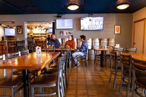 Three people sit at a table in a casual restaurant, with kegs in the background and screens displaying menus.