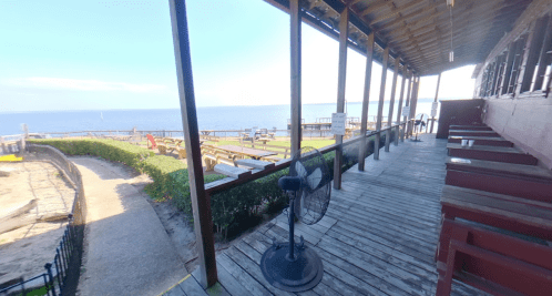 View of a wooden deck by the water, featuring a fan and seating, with a sandy area and greenery in the background.