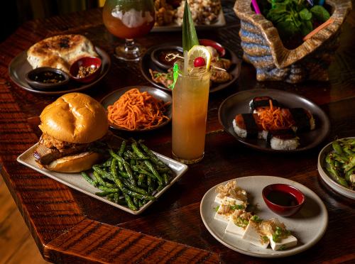 A table spread with a burger, green beans, sushi, noodles, and a colorful cocktail, surrounded by various dishes.