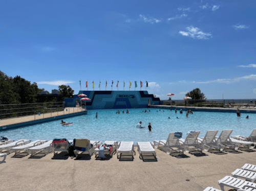 A sunny outdoor pool with people swimming, lounge chairs, and colorful flags in the background.