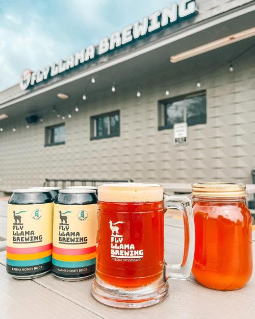 Three cans and a jar of beer in front of Fly Llama Brewing, with a glass of amber beer on a wooden table.