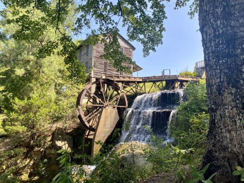 A historic watermill with a large wooden wheel, surrounded by lush greenery and a flowing waterfall.