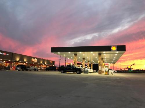 A Shell gas station at sunset, with colorful skies and vehicles parked at the pumps. Nearby shops are visible.
