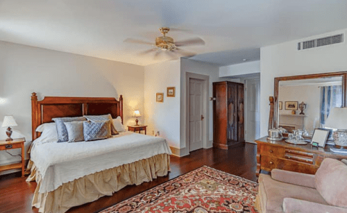 Cozy bedroom with a wooden bed, soft lighting, and a vintage rug, featuring a dresser and a comfortable armchair.