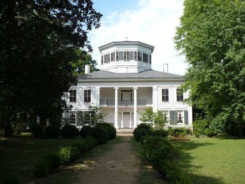 A large white historic house with a circular tower, surrounded by greenery and a pathway leading to the entrance.